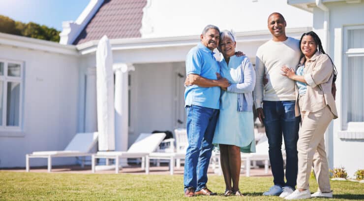 A multigenerational family of four stands on a lawn in front of a white house, smiling and embracing.