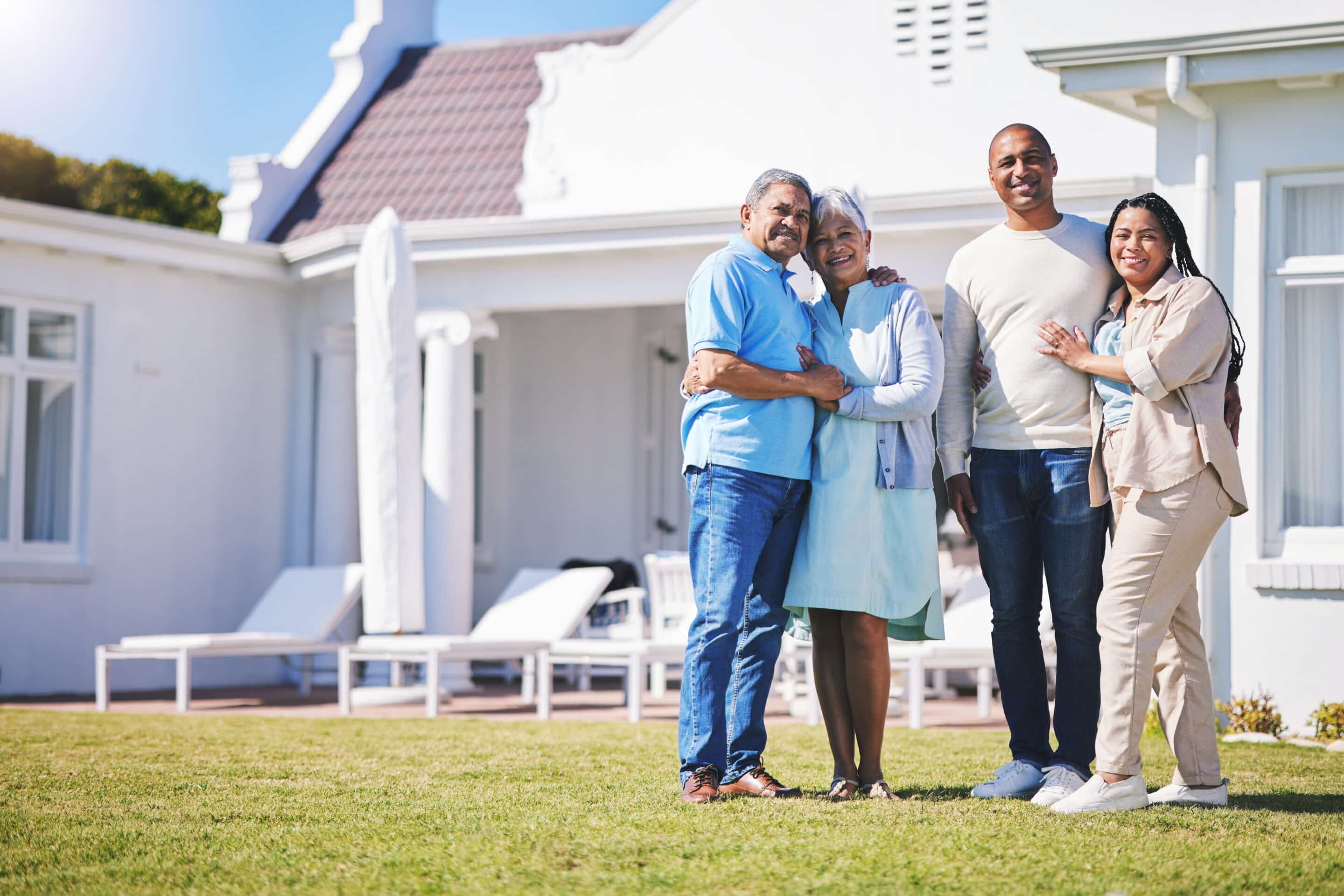 A multigenerational family of four stands on a lawn in front of a white house, smiling and embracing.