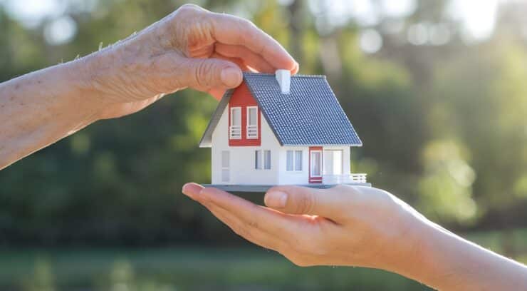 Two hands pass a miniature house between them outdoors, symbolizing a real estate handover or gift.