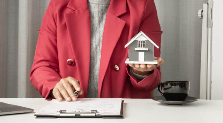 Person in a red blazer presents a small gray house model while seated at a desk with papers and a coffee cup nearby.