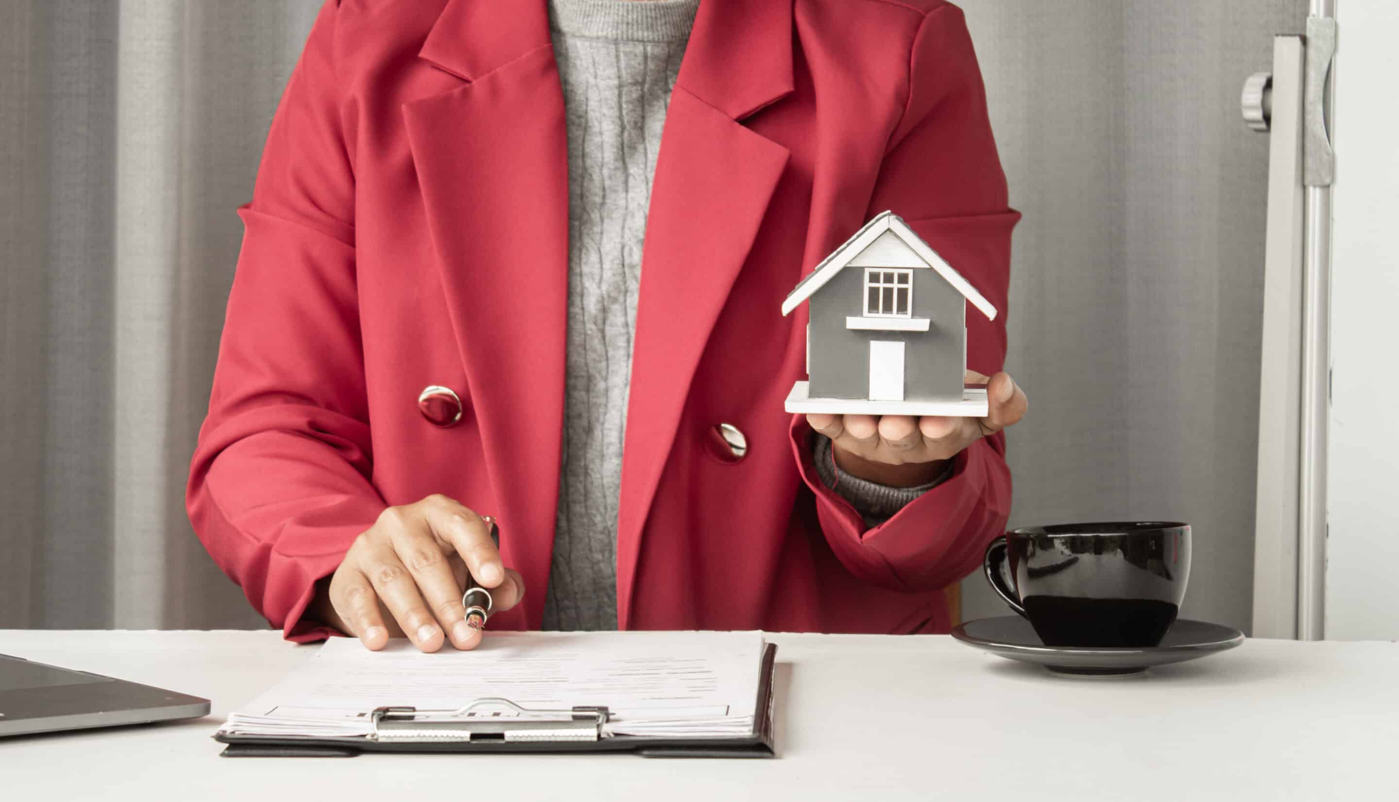 Person in a red blazer presents a small gray house model while seated at a desk with papers and a coffee cup nearby.
